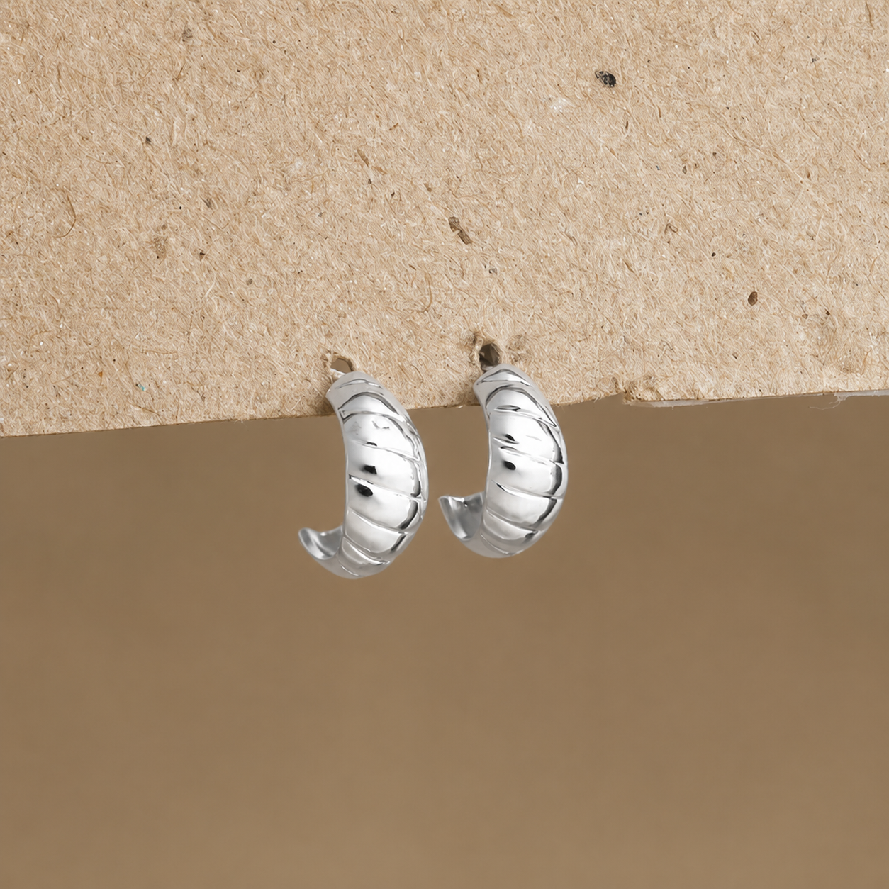 A pair of small silver hoop earrings with a textured, ribbed outer surface, displayed against a split beige and brown background.